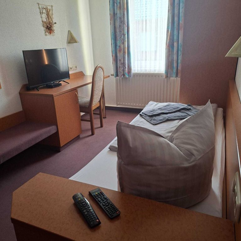 Schlafzimmer mit Bett, Schreibtisch, Stuhl und Fernseher. Tageslicht durch das Fenster.